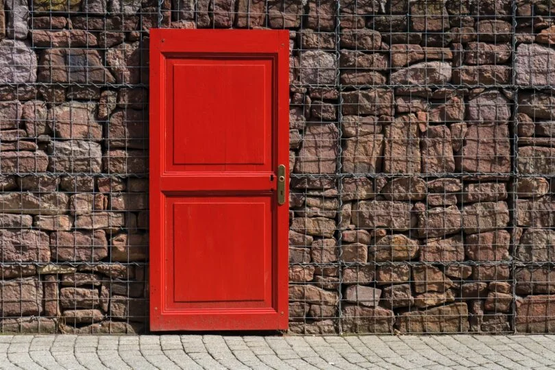 Red door in front of rocks held by fencing. 